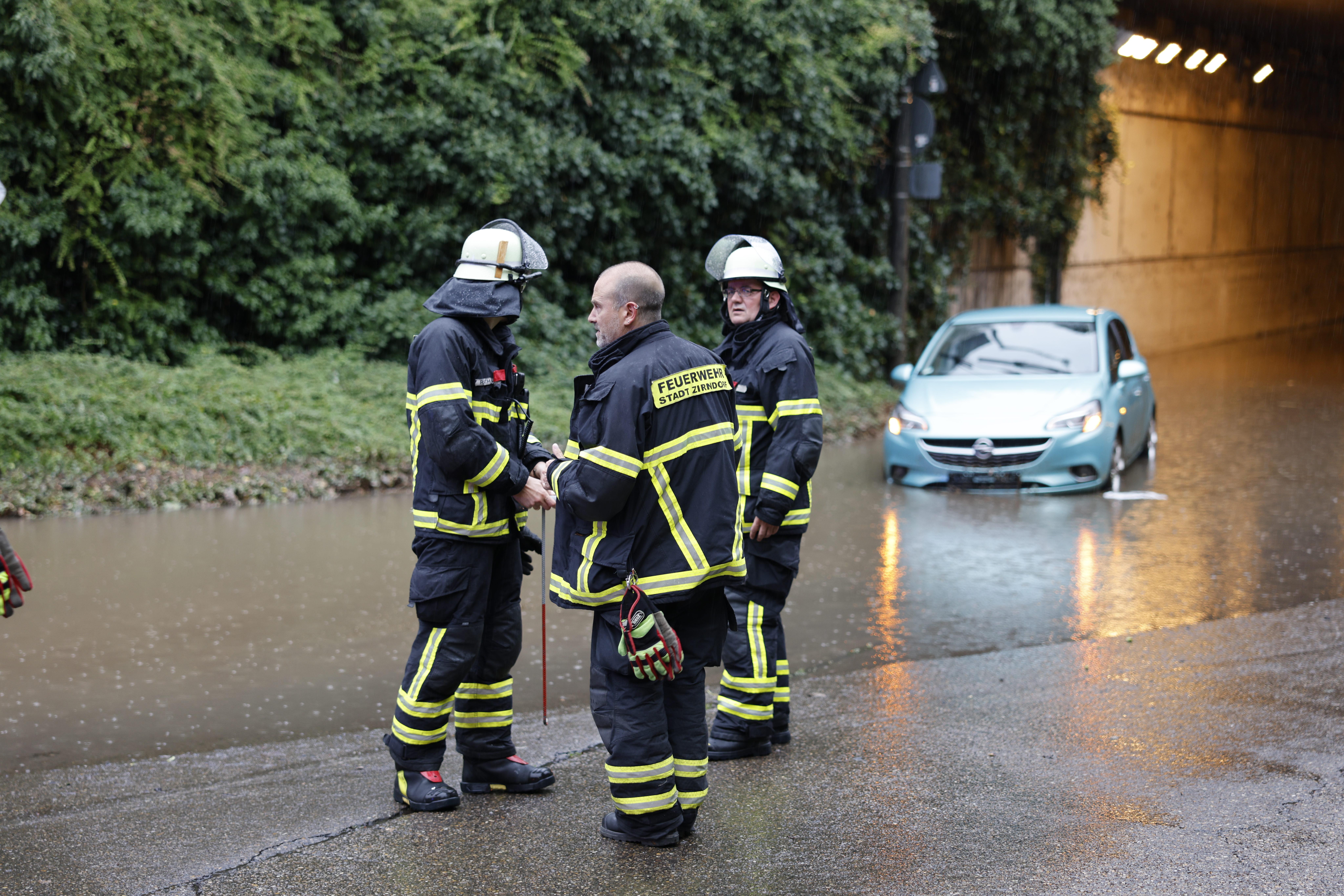 Drei Feuerwehrleute stehen neben einer überfluteten Unterführung, im Hintergrund ein Auto im Wasser.