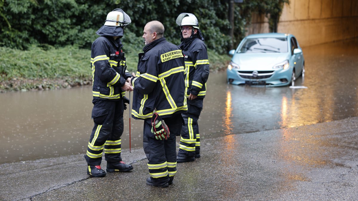 Drei Feuerwehrleute stehen neben einer überfluteten Unterführung, im Hintergrund ein Auto im Wasser.