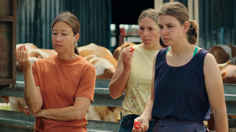 Szene aus dem Film "Milch ins Feuer". Eine von Johanna Wokalek gespielte Milchbäuerin steht mit zwei ihrer Töchter zusammen und isst Tomaten. | Bild: Bauer Carnicer / Filmperlen Szene aus dem Film "Milch ins Feuer". Eine von Johanna Wokalek gespielte Milchbäuerin steht mit zwei ihrer Töchter zusammen und isst Tomaten.