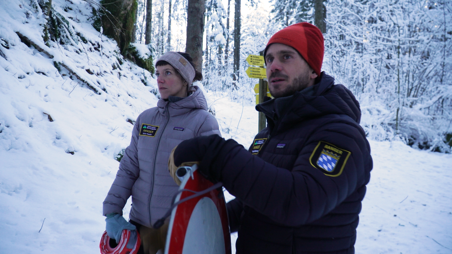 Eine Frau und ein Mann stehen im Bergwald und halten ein Absperrband und ein Stopp-Schild in den Händen