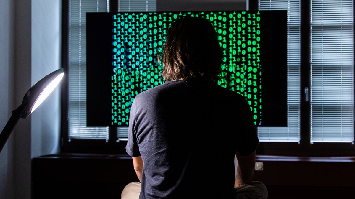 Ein Mann sitzt vor einem Bildschirm, auf dem ein Binärcode läuft.