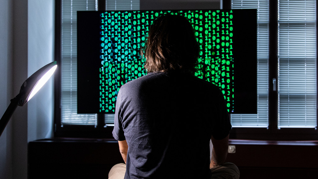 Ein Mann sitzt vor einem Bildschirm, auf dem ein Binärcode läuft.
