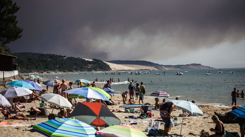 Frankreich, Pyla Sur Mer: Strandbesucher baden, während eine schwarze Rauchwolke von einem Feuer im Hintergrund aufsteigt.