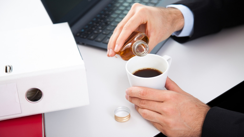 Illustration - Ein Mann gießt Alkohol in seinen Kaffee am Arbeitsplatz | Bild: picture alliance / dpa Themendienst | Christin Klose Illustration - Ein Mann gießt Alkohol in seinen Kaffee am Arbeitsplatz