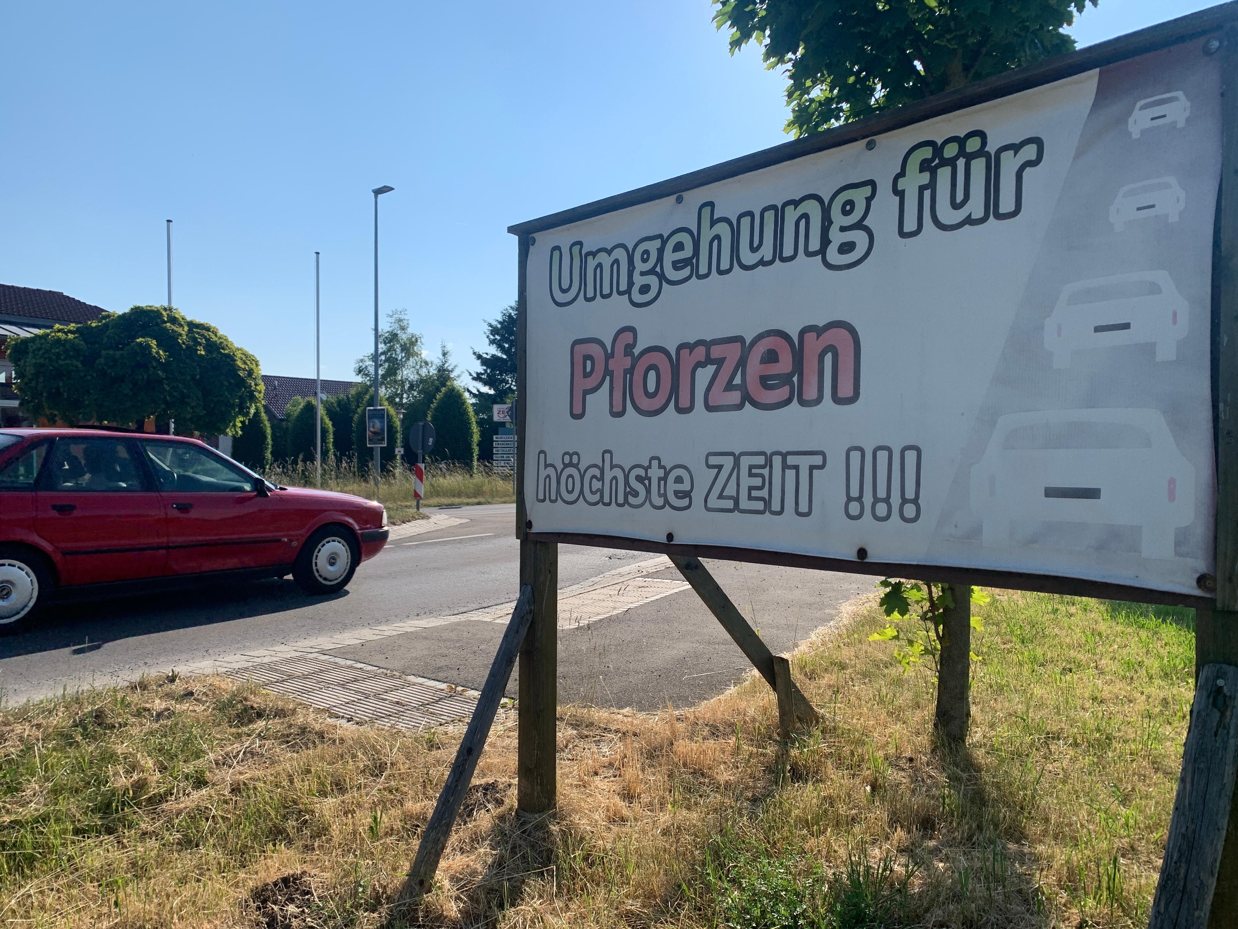 "Umgehung für Pforzen höchste Zeit!!!" steht auf einem Plakat, das am Ortseingang von Pforzen aufgestellt wurde. Gerade fährt ein rotes Auto vorbei. Seit gut zehn Jahren wird in Pforzen um eine Ortsumfahrung gerungen (Bild von Juni 2023).