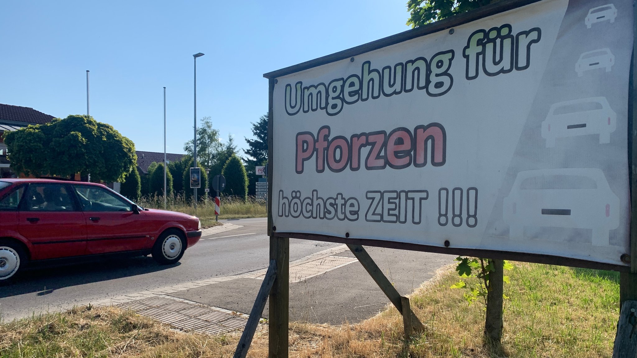 "Umgehung für Pforzen höchste Zeit!!!" steht auf einem Plakat, das am Ortseingang von Pforzen aufgestellt wurde. Gerade fährt ein rotes Auto vorbei. Seit gut zehn Jahren wird in Pforzen um eine Ortsumfahrung gerungen (Bild von Juni 2023).