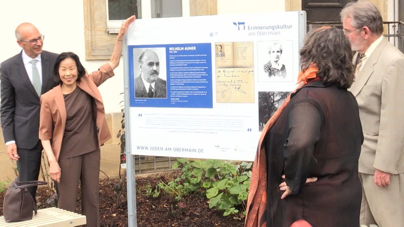Zwei Männer und zwei Frauen stehen vor einer Tafel, die ein Foto von Wilhelm Aumer zeigt. Dieser hatte während der NS-Diktatur jüdischen Mitbürgern die Ausreise ermöglicht.