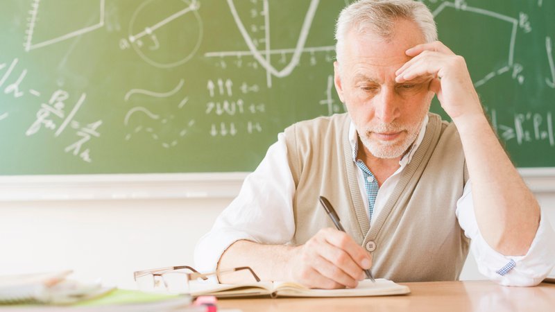 Älterer Lehrer sitzt müde am Pult vor Tafel | Bild: picture alliance / imageBROKER | Oleksandr Latkun Älterer Lehrer sitzt müde am Pult vor Tafel