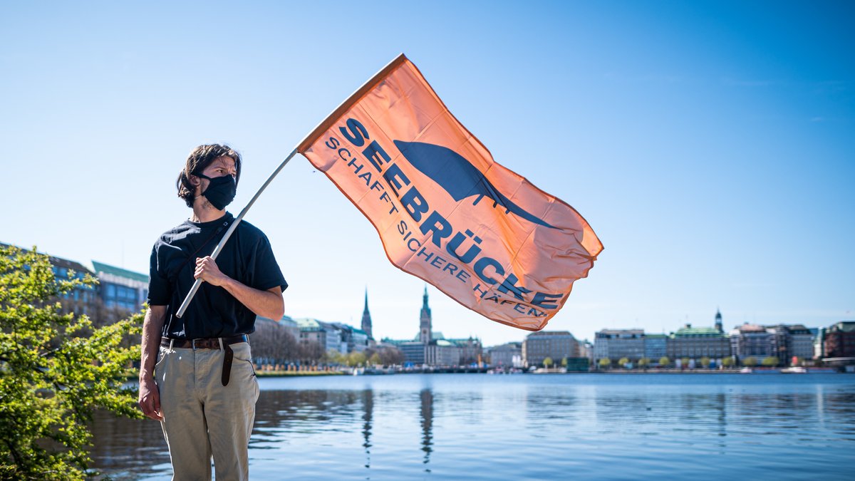 Mann vor Wasser mit Flagge in der Hand mit dem Titel: Seebrücke Schafft sichere Häfen 