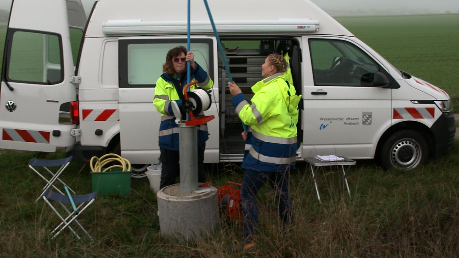 Zwei Frauen in Arbeitskleidung stecken ein dünnes blaues Rohr in das dicke Rohr der Messstelle. Dahinter ihr Dienstauto.