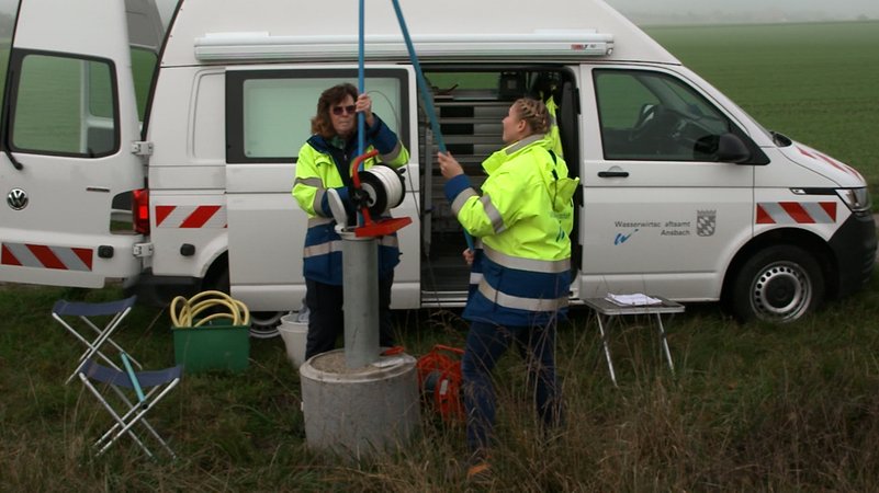 Zwei Frauen in Arbeitskleidung stecken ein dünnes blaues Rohr in das dicke Rohr der Messstelle. Dahinter ihr Dienstauto. | Bild: BR / David Sünderhauf Zwei Frauen in Arbeitskleidung stecken ein dünnes blaues Rohr in das dicke Rohr der Messstelle. Dahinter ihr Dienstauto.