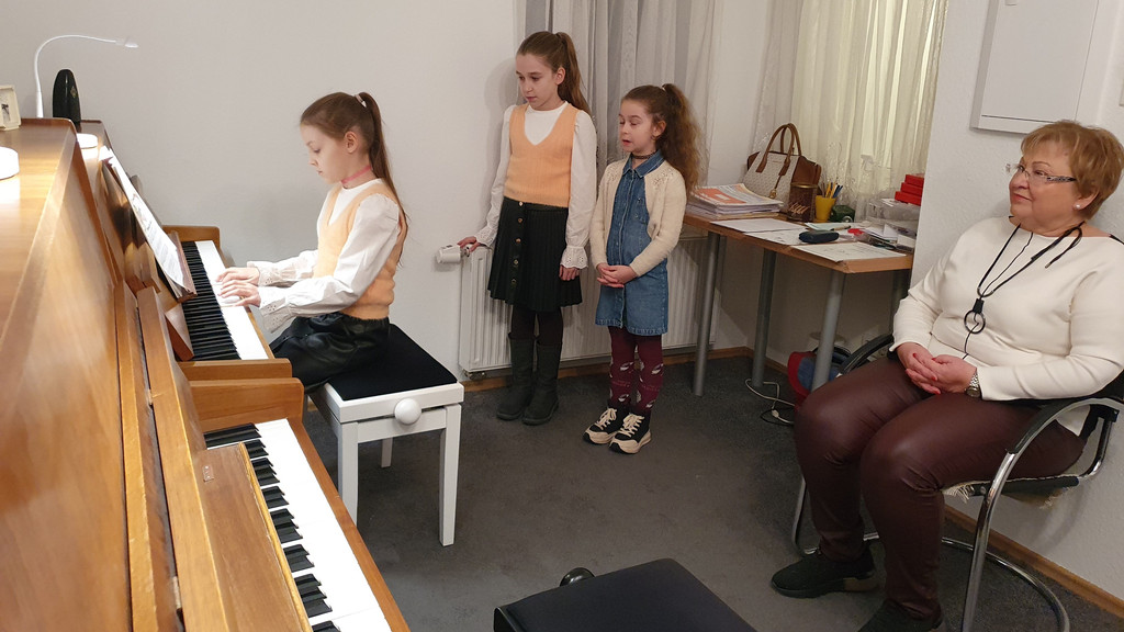 Ein Mädchen sitzt am Klavier und spielt, seine beiden Schwestern stehen hinter ihr und singen.