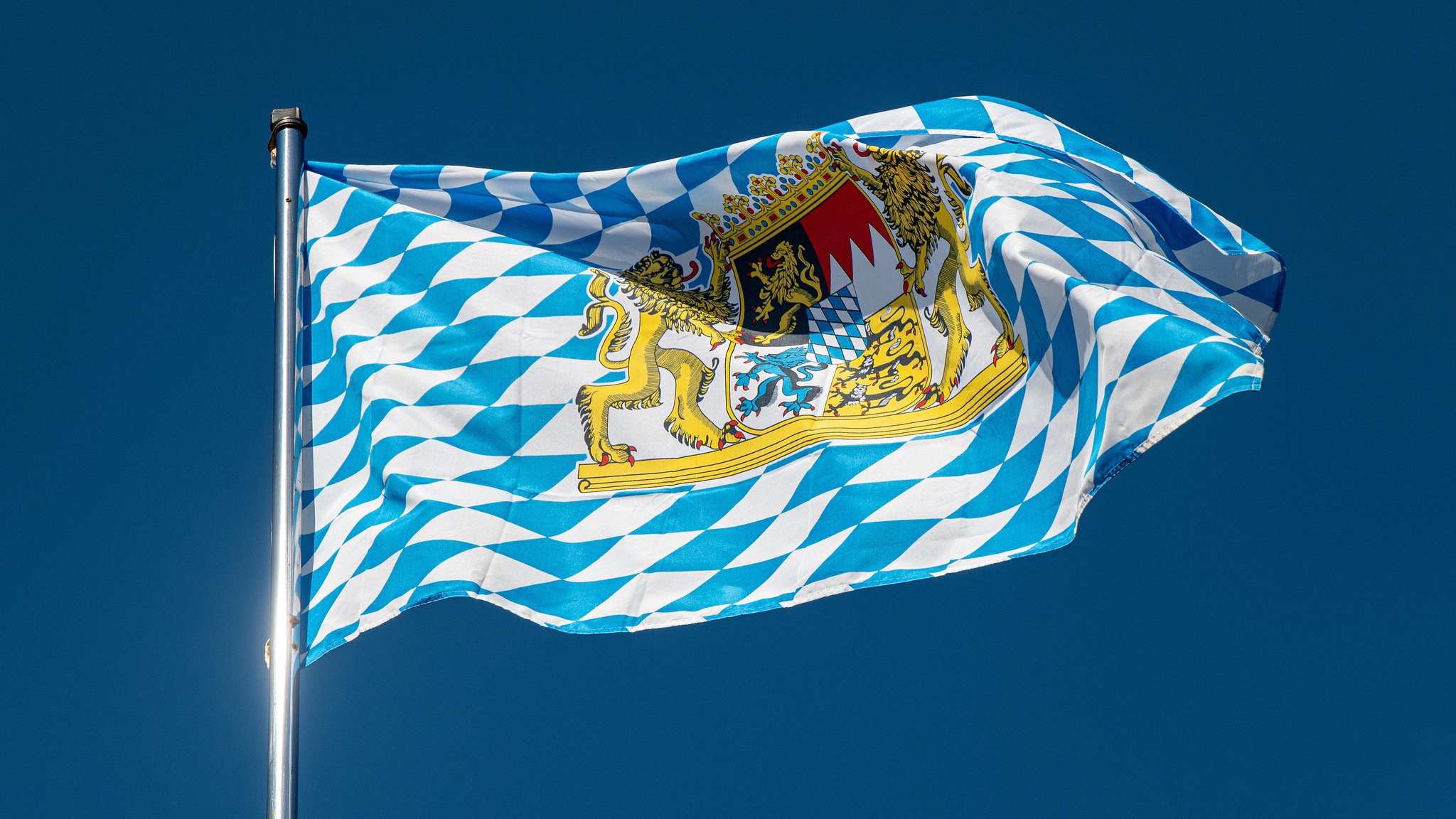 Die bayerische Landesflagge mit dem großen Staatswappen und den typischen weiß-blauen Rauten vor blauem Himmel.