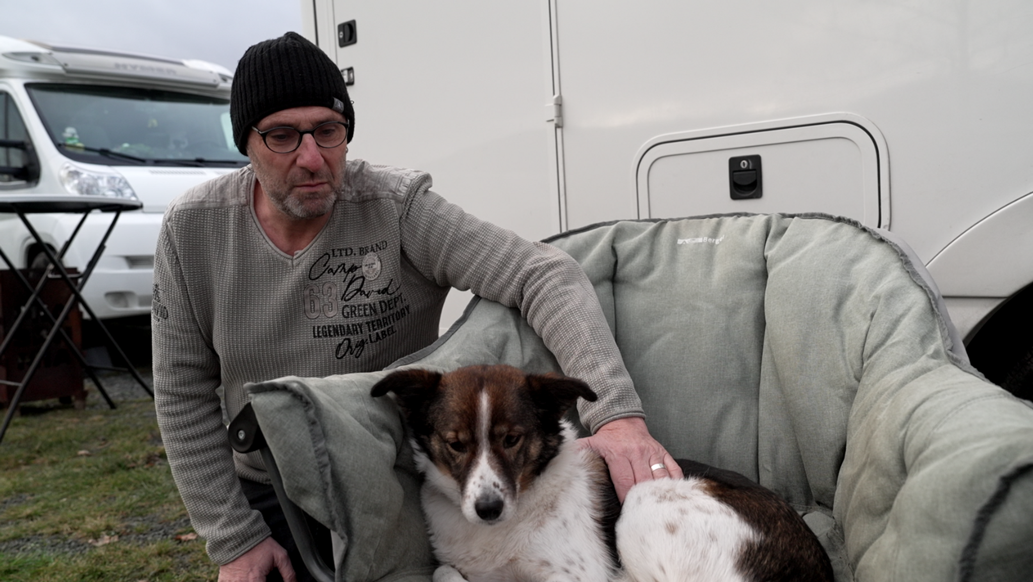 Ein Mann mit grauem Pullover und schwarzer Mütze kniet neben einem Stuhl. Auf dem Stuhl liegt ein weißer Hund mit braunem Kopf. 