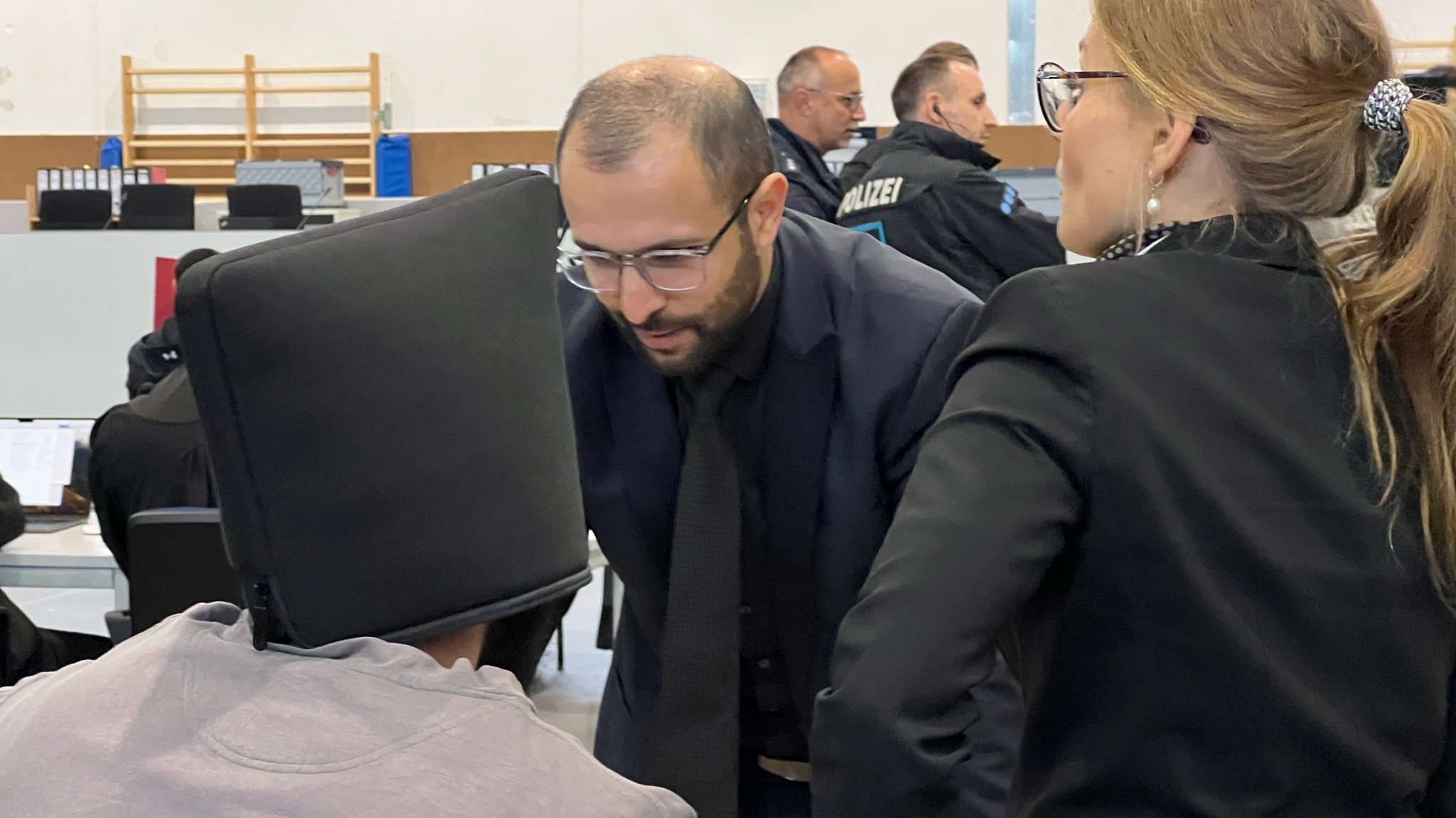 Ein Mann, der eine Tasche auf seinem Kopf trägt, im Gespräch mit seinem Anwalt. Im Hintergrund stehen Polizisten. 