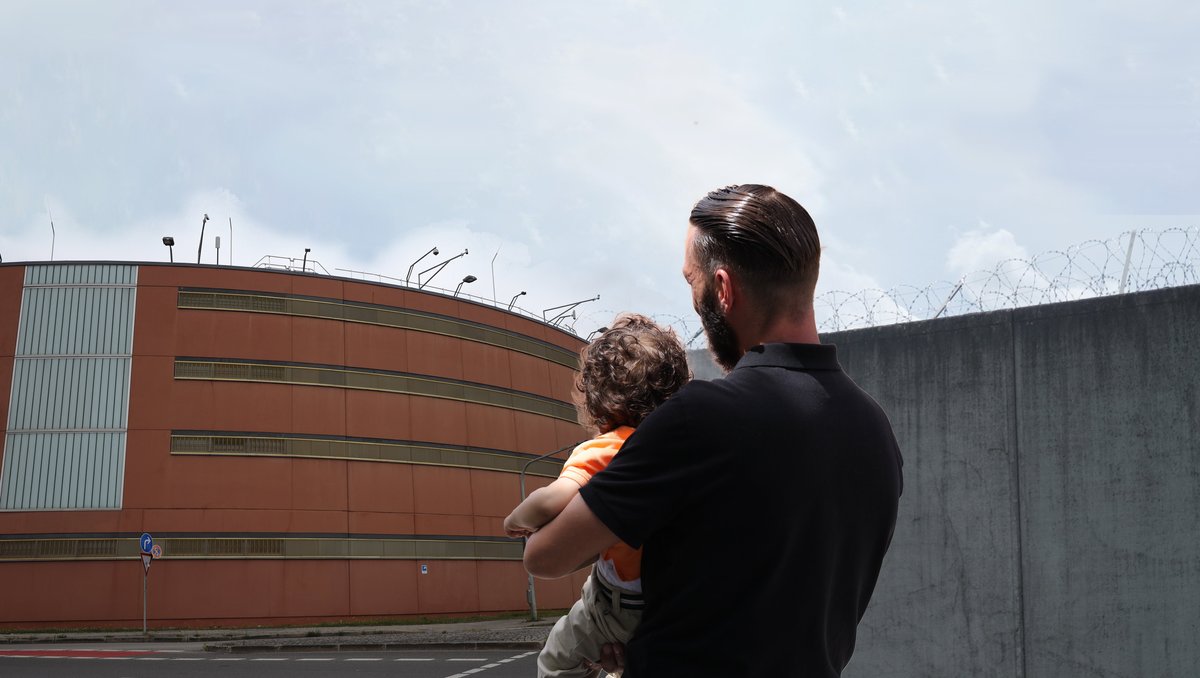Ein Mann mit Kind auf dem Arm steht vor einem Gefängnis. 