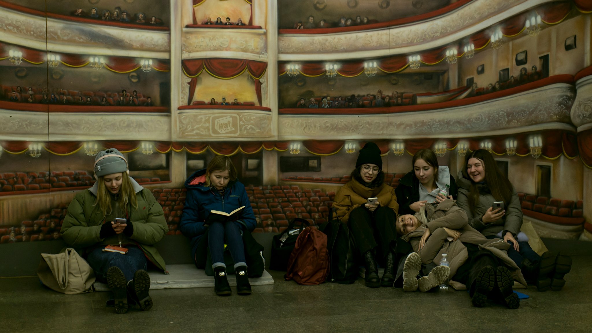 Junge Menschen sitzen in einer U-Bahn-Station in Kiew auf dem Boden, lesen und schauen auf ihre Smartphones.