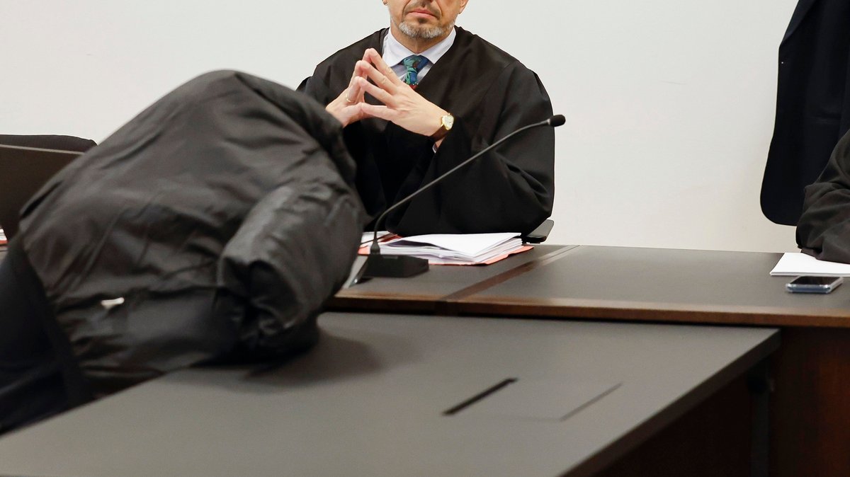 Ein Angeklagter verdeckt seinen Kopf mit einer schwarzen Jacke, sein Verteidiger sitzt im Hintergrund.