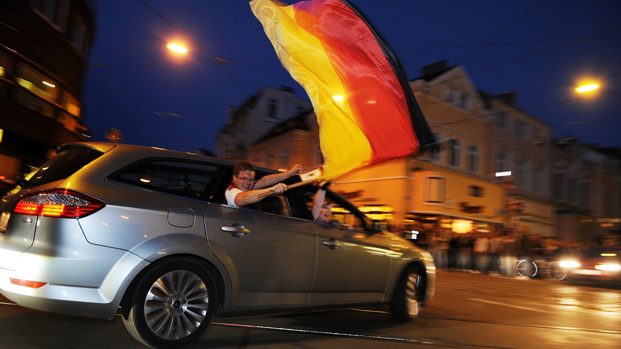 Fußball-Fans fahren mit einem Auto durch eine Innenstadt. Ein Mann schwenkt eine Fahne.