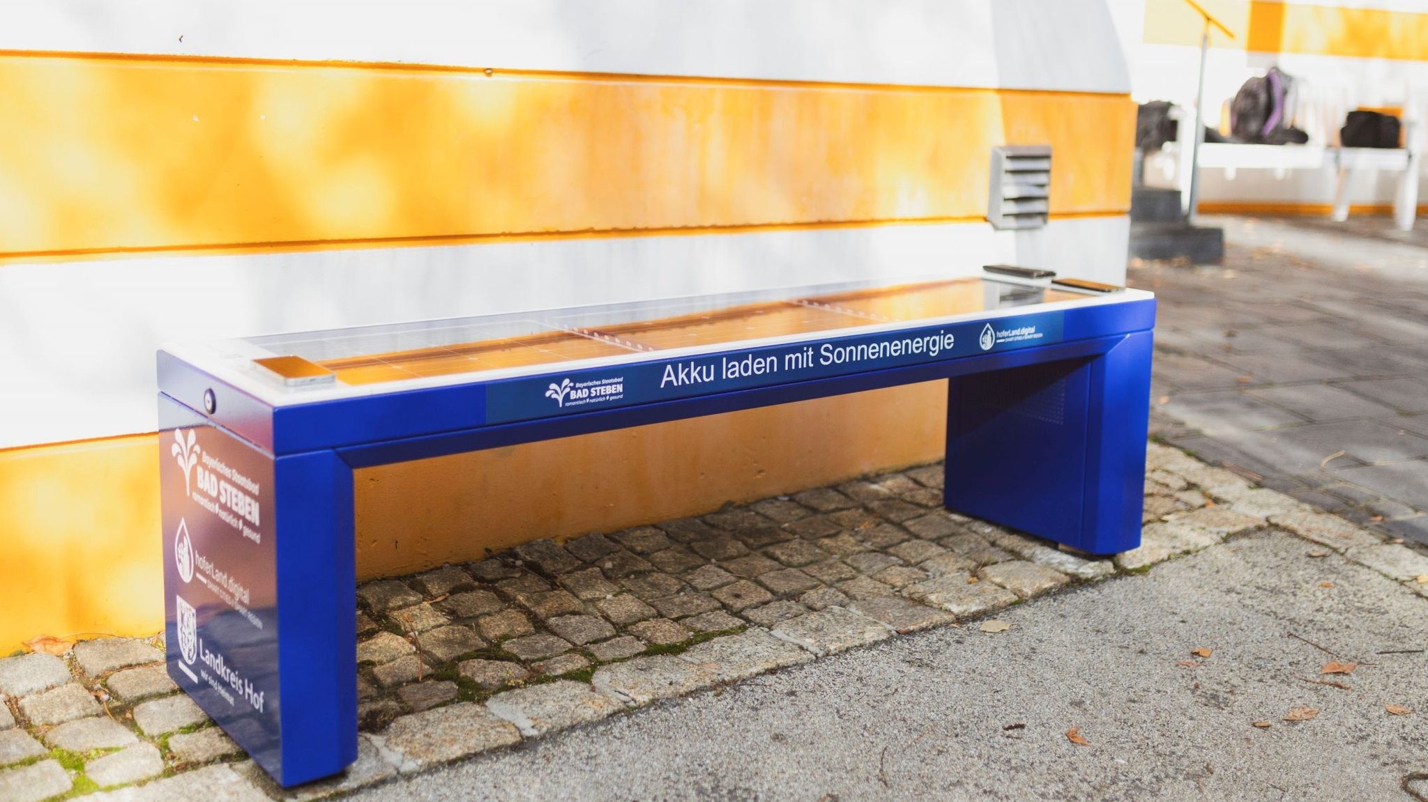 Eine blaue Bank ohne Lehne steht vor einer weiß-gelb gestreiften Wand.