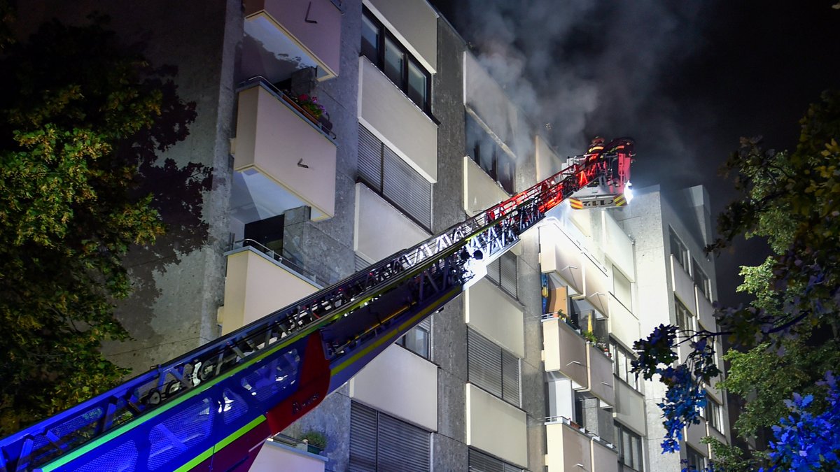 Einsatzkräfte der Münchner Feuerwehr löschen das Feuer im vierten Stock von einer Drehleiter aus; starker Rauch quillt aus einem der Fenster.