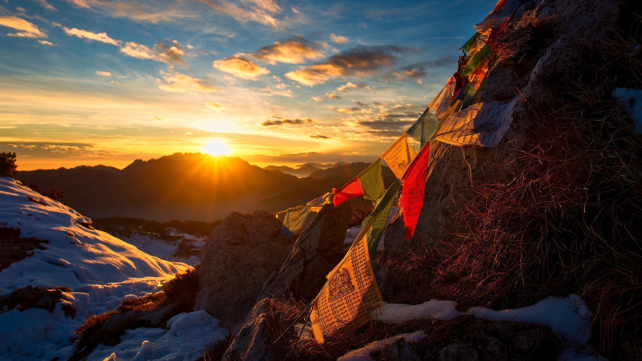 Tibetische Gebetsfahnen im Vordergrund, ein Sonnenuntergang in den Bergen im Hintergrund. | Bild: picture alliance / CHROMORANGE | Michelangelo Oprandi Tibetische Gebetsfahnen im Vordergrund, ein Sonnenuntergang in den Bergen im Hintergrund.