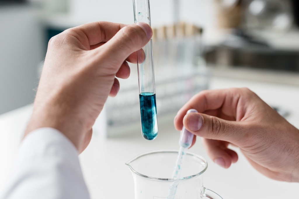 Symbolbild Labor: Eine Person hält mit einer Hand ein Reagenzglas, mit dem anderen eine Pipette. Blau gefärbte Flüssigkeit wird pipettiert.
