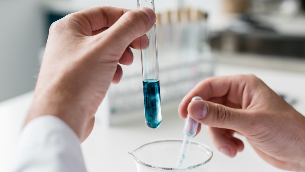 Symbolbild Labor: Eine Person hält mit einer Hand ein Reagenzglas, mit dem anderen eine Pipette. Blau gefärbte Flüssigkeit wird pipettiert.