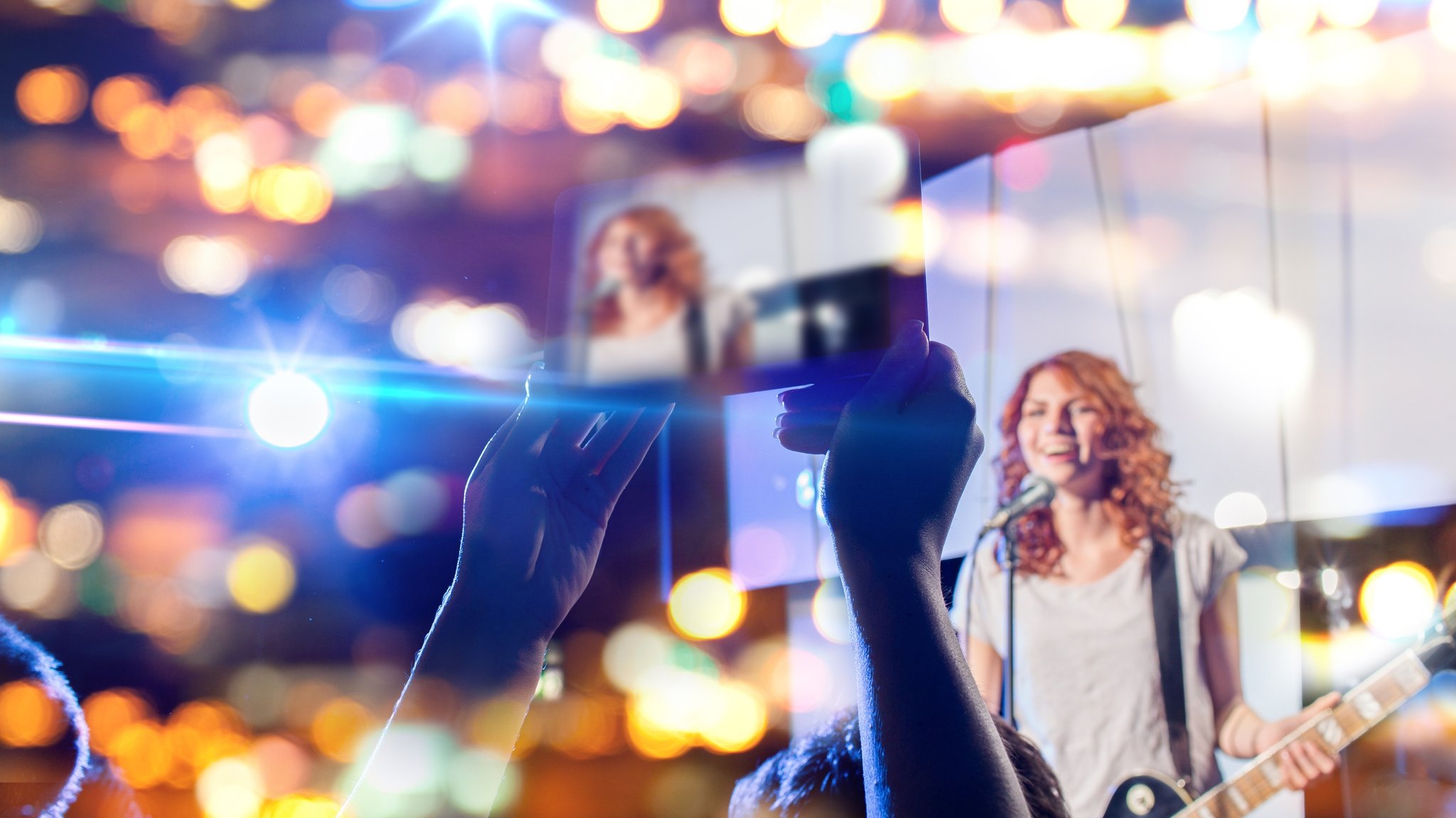 Bei einem Livekonzert wird eine Sängerin, die E-Gitarre spielt und auf der Bühne singt, von einem Fan mit dem Handy aufgenommen.