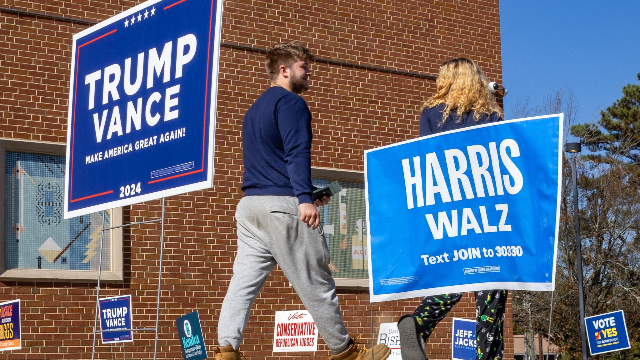 Zwei Menschen gehen zum Wahllokal, neben ihnen Schilder von Harris und Trump.