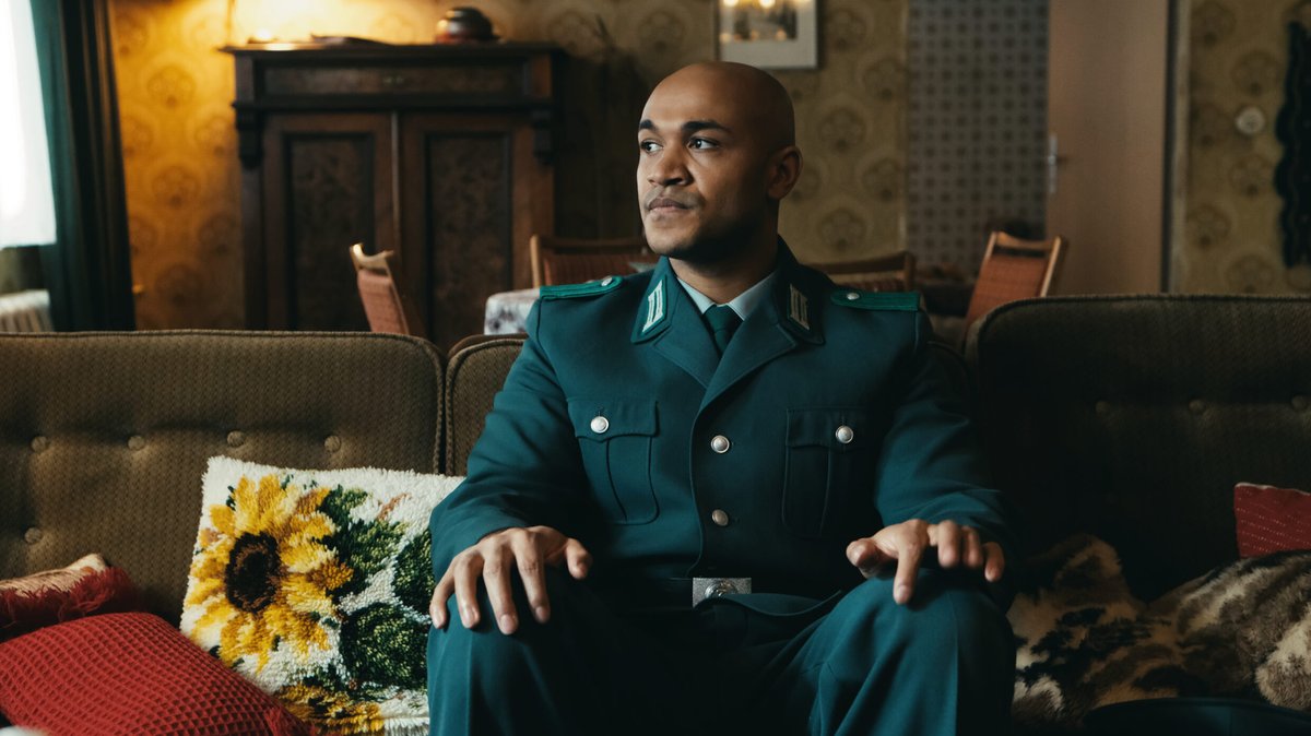 Sam Meffire (Malick Bauer) war der erste afrodeutsche Polizist in Ostdeutschland. Er sitzt uniformiert auf einem Sofa und schaut an der Kamera vorbei.