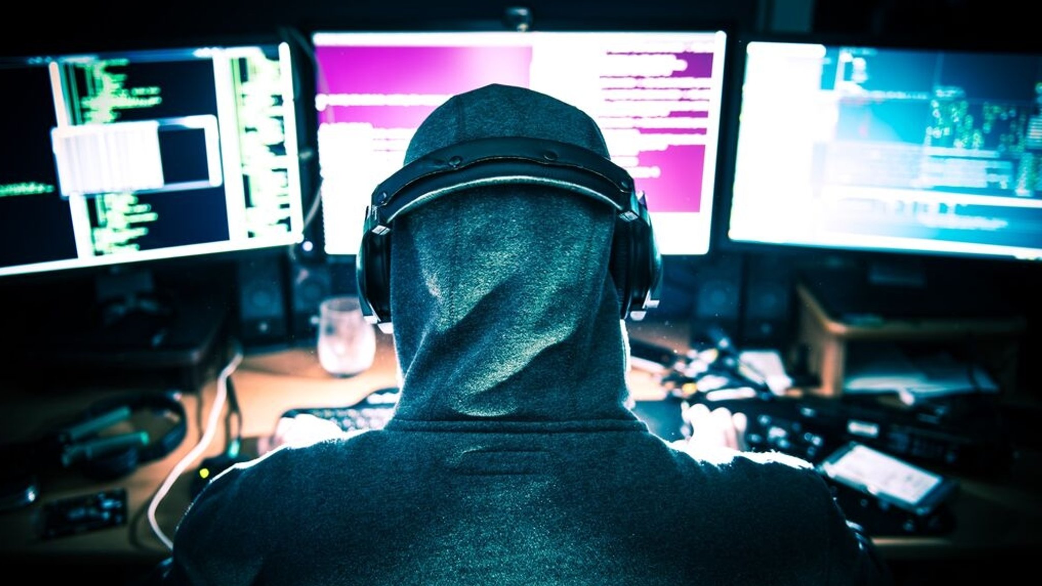 Eine Person mit Kapuzenpullover und Kopfhörern sitzt vor mehreren Monitoren, die mit Code und Daten gefüllt sind (Symbolbild)