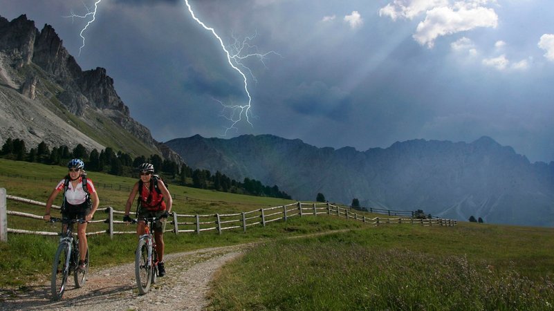 Zwei Fahrradfahrerinnen bei Blitz unterwegs in den Bergen | Bild: picture alliance / imageBROKER | Joerg Reuther Zwei Fahrradfahrerinnen bei Blitz unterwegs in den Bergen