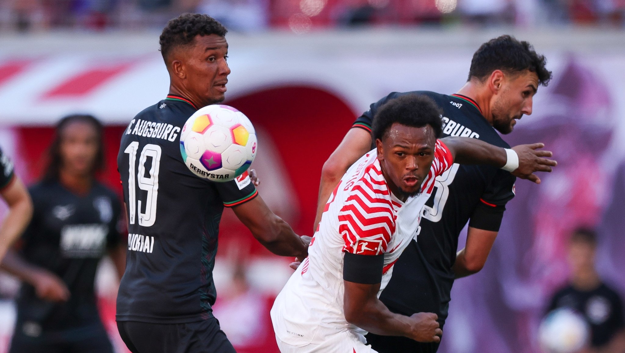 16.09.2023, Sachsen, Leipzig: Fußball: Bundesliga, 4. Spieltag, RB Leipzig - FC Augsburg in der Red-Bull-Arena. Leipzigs Lois Openda (M) im Duell mit Augsburgs Felix Uduokhai (l) und Maximilian Bauer. 