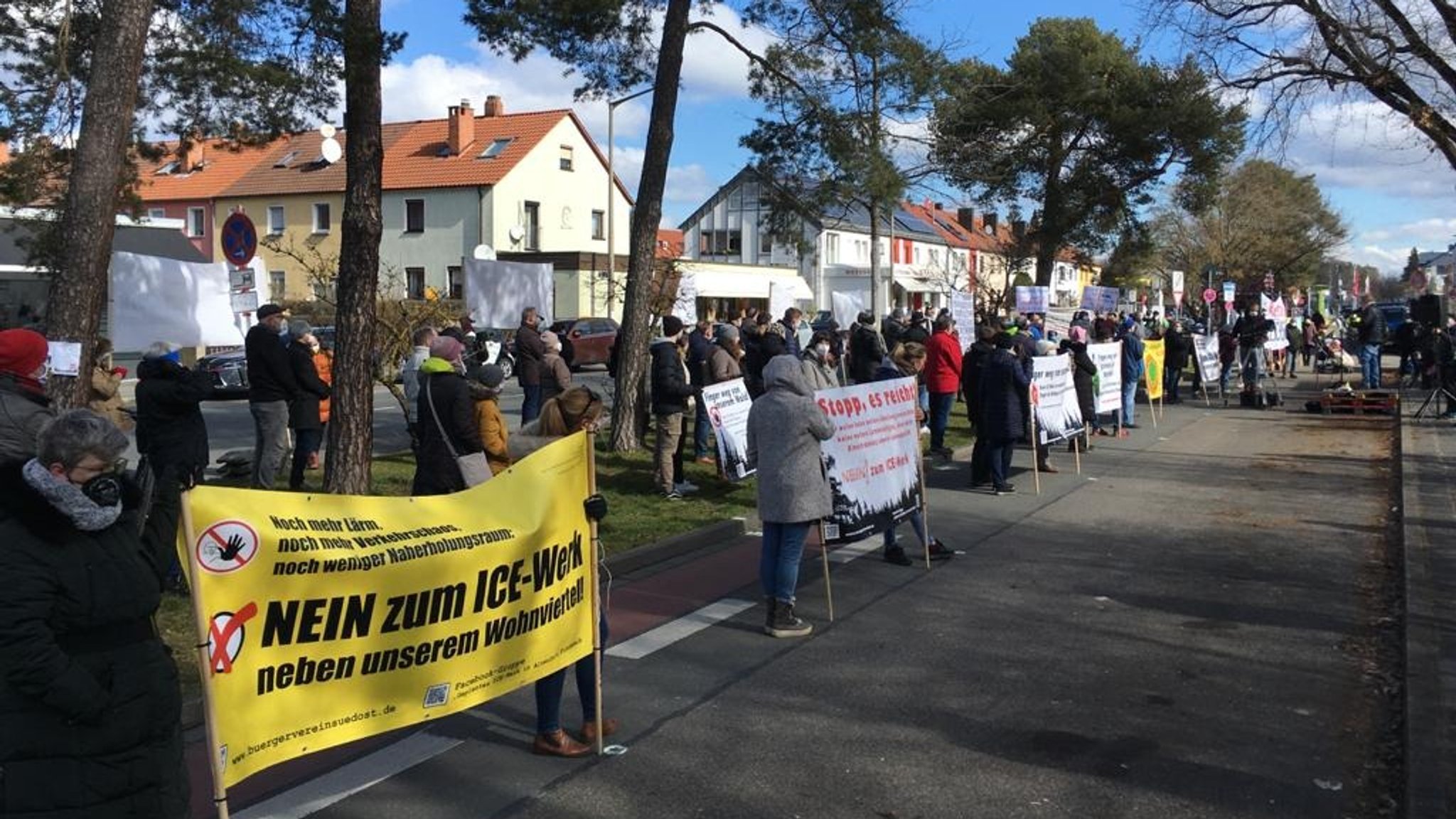 Menschen mit Transparenten "Nein zum ICE-Werk neben unserem Wohnviertel" und anderen in einer Allee.