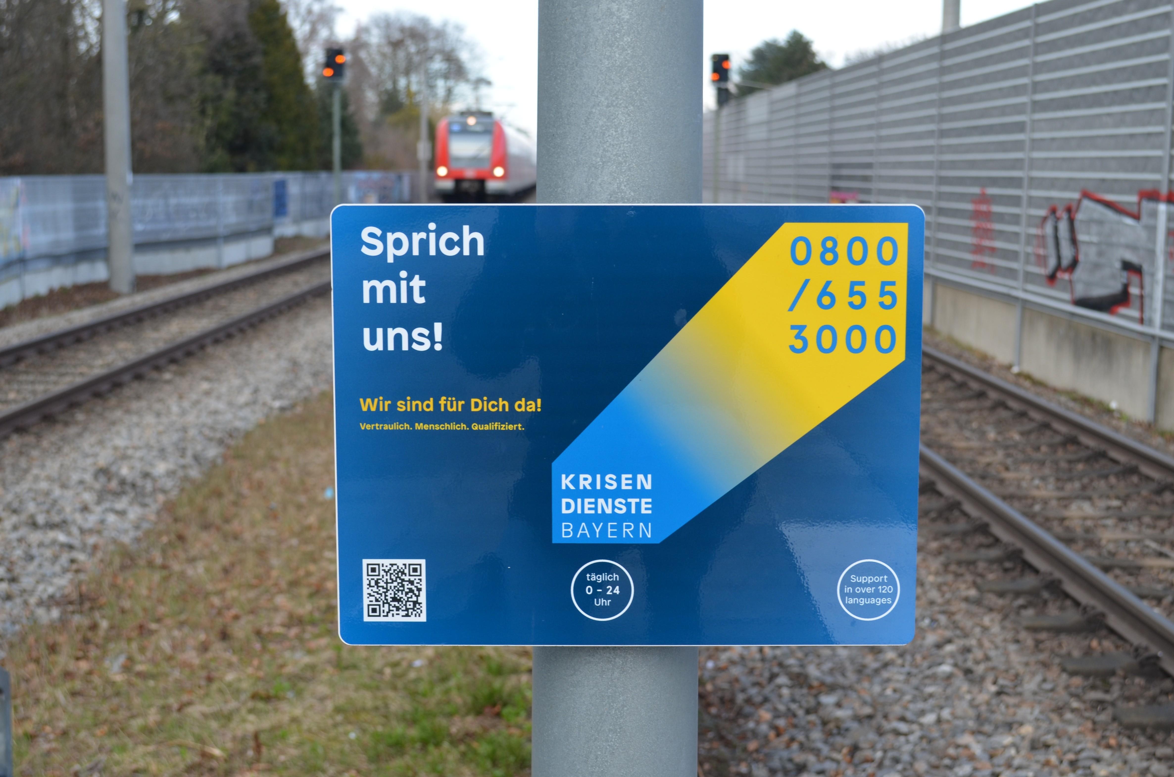 Ein Schild der Krisendienste an einem Bahngleis mit einfahrendem Zug