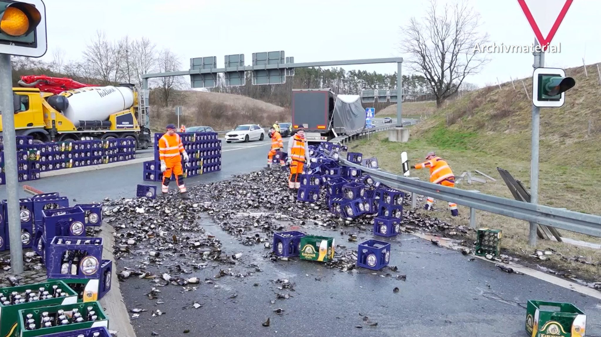 Bierkästen und zerborstene Bierflaschen liegen auf der Straße. Männer in orangenen Anzügen stehen daneben. Im Hintergrund sind stehende Autos, ein Lkw und ein Betonmischer zu sehen.