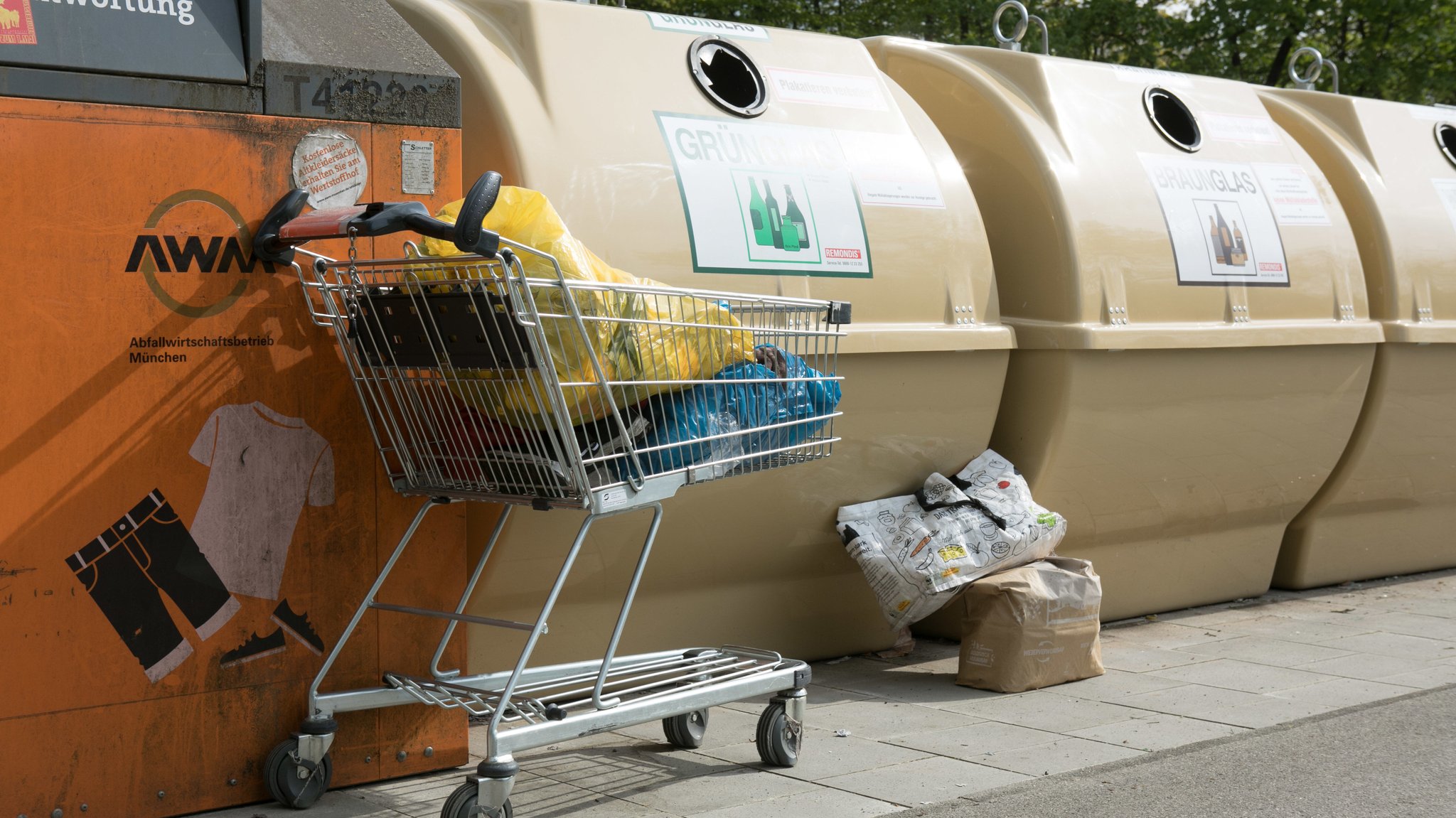 (Symbolbild) Ein mit Müll gefüllter Einkaufswage steht vor einer Wertstoffinsel