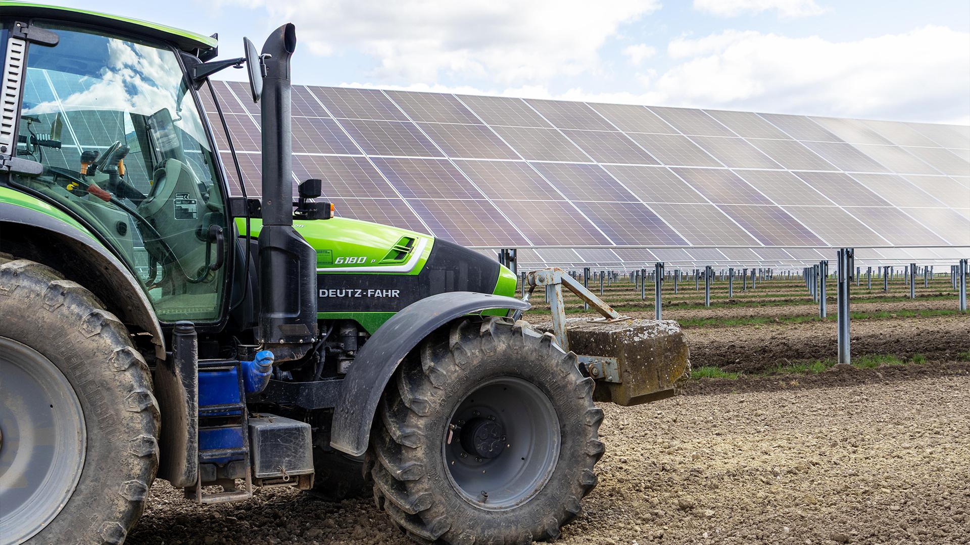 Deutz Traktor steht auf einem Feld vor einer großflächigen Agrivoltaik-Anlage mit geneigten Solarmodulen.