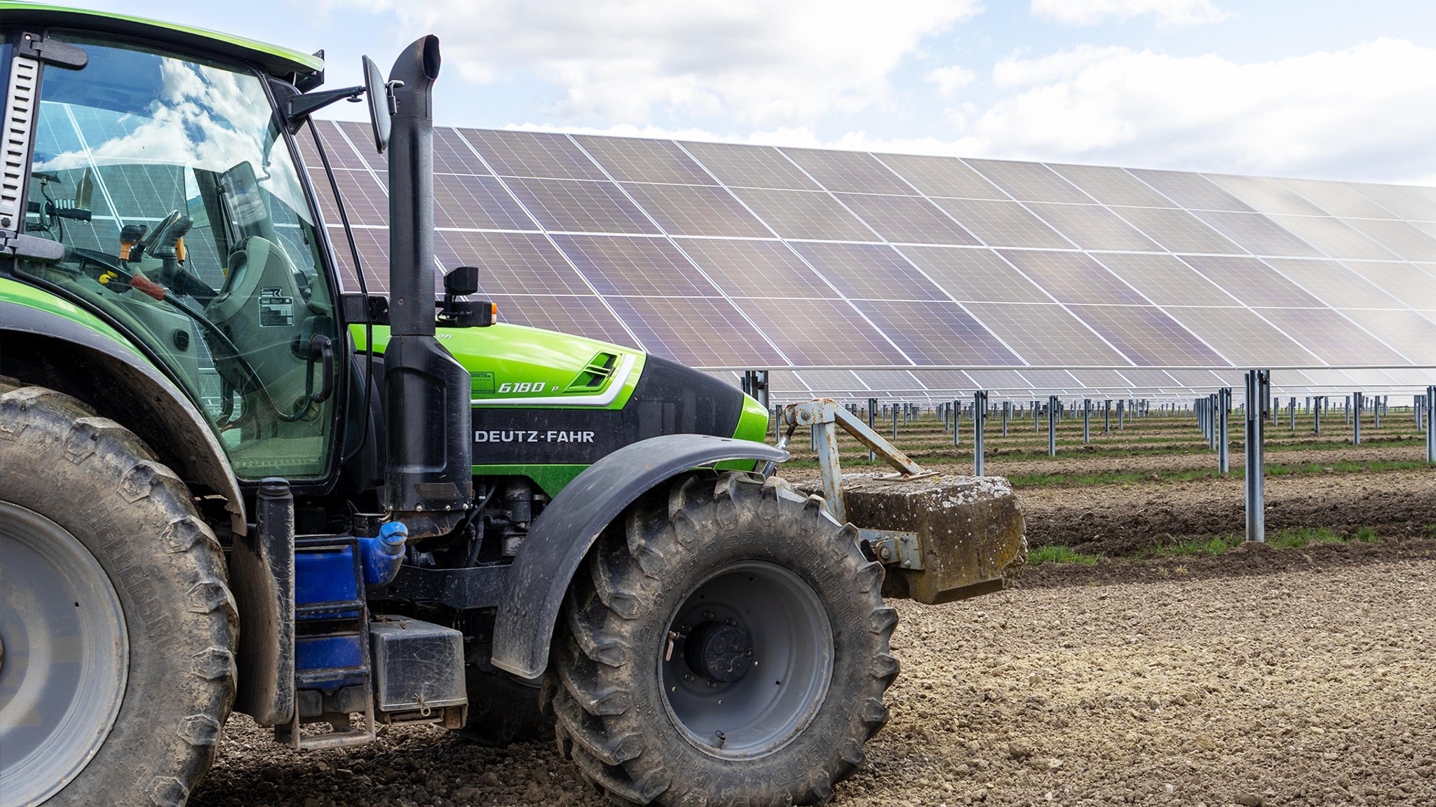 Deutz Traktor steht auf einem Feld vor einer großflächigen Agrivoltaik-Anlage mit geneigten Solarmodulen.