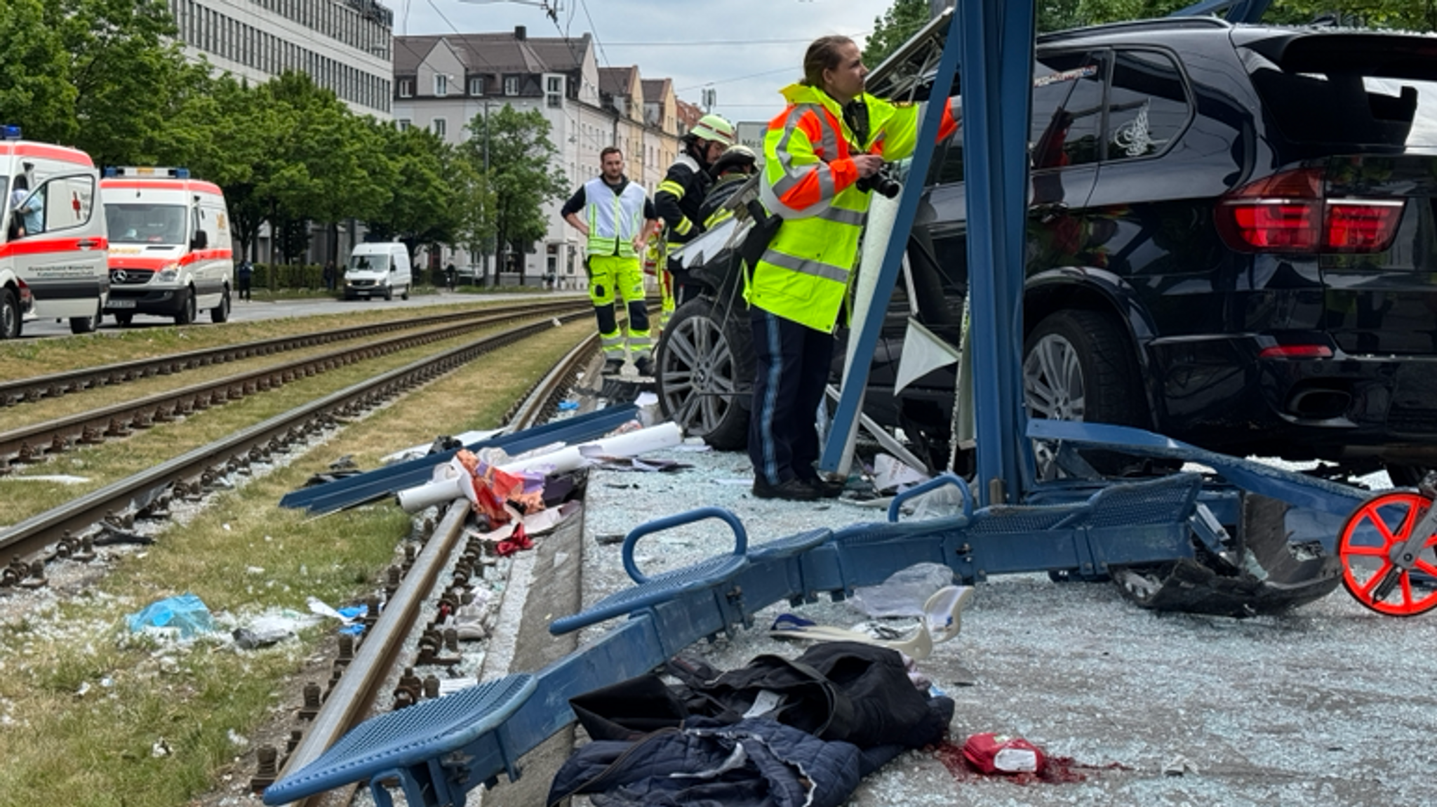 07.05.2025, München: Polizisten und Rettungskräfte stehen neben einem Unfallfahrzeug in den Glassplittern einer Haltestelle.