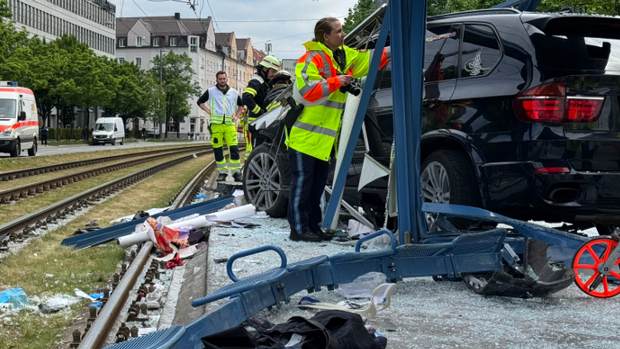 07.05.2025, München: Polizisten und Rettungskräfte stehen neben einem Unfallfahrzeug in den Glassplittern einer Haltestelle.