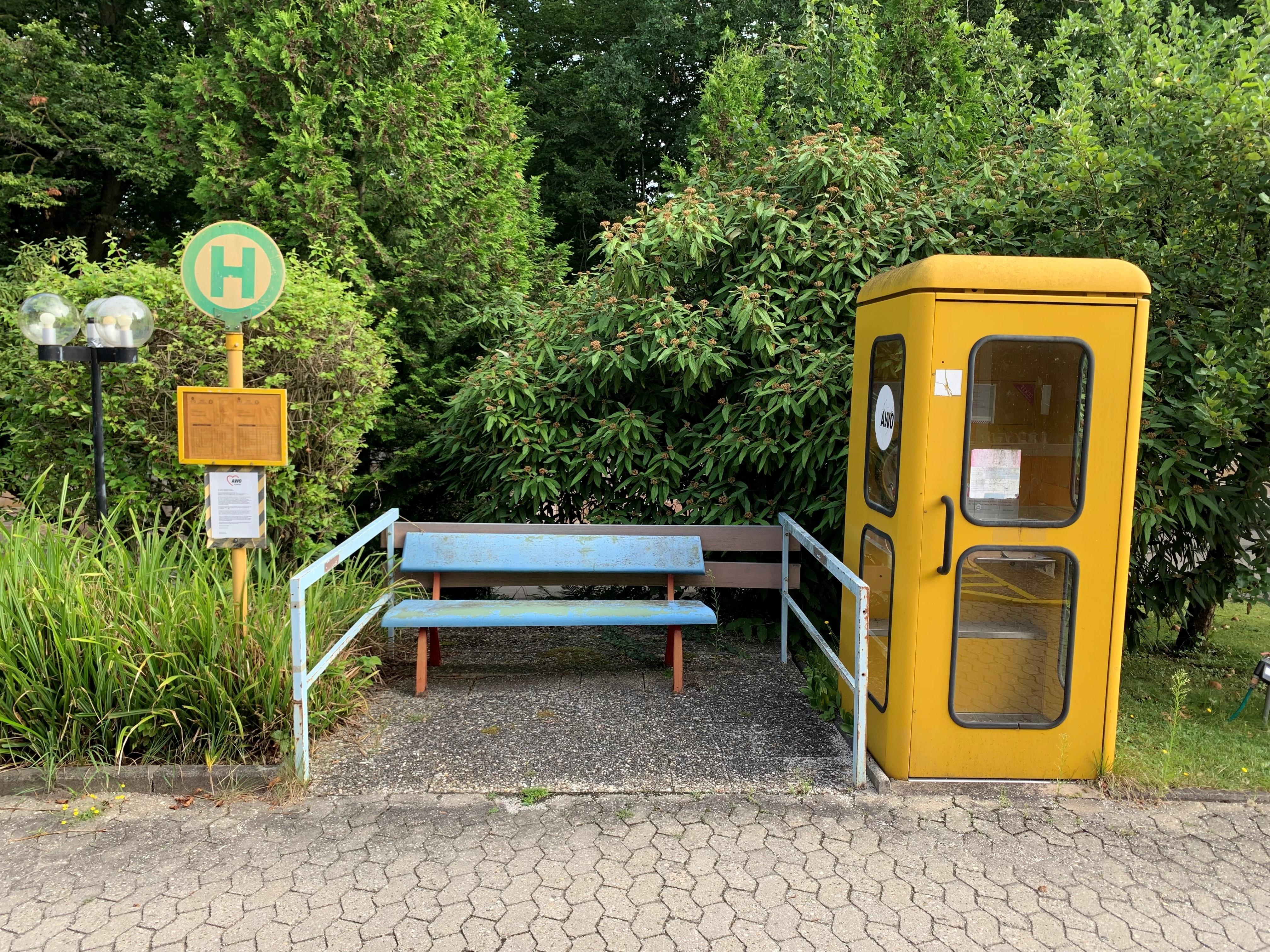 Neben einem Haltestellenschild stehen eine Bank und eine Telefonzelle. 