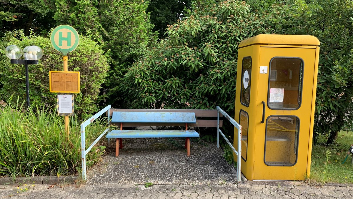 Neben einem Haltestellenschild stehen eine Bank und eine Telefonzelle. 