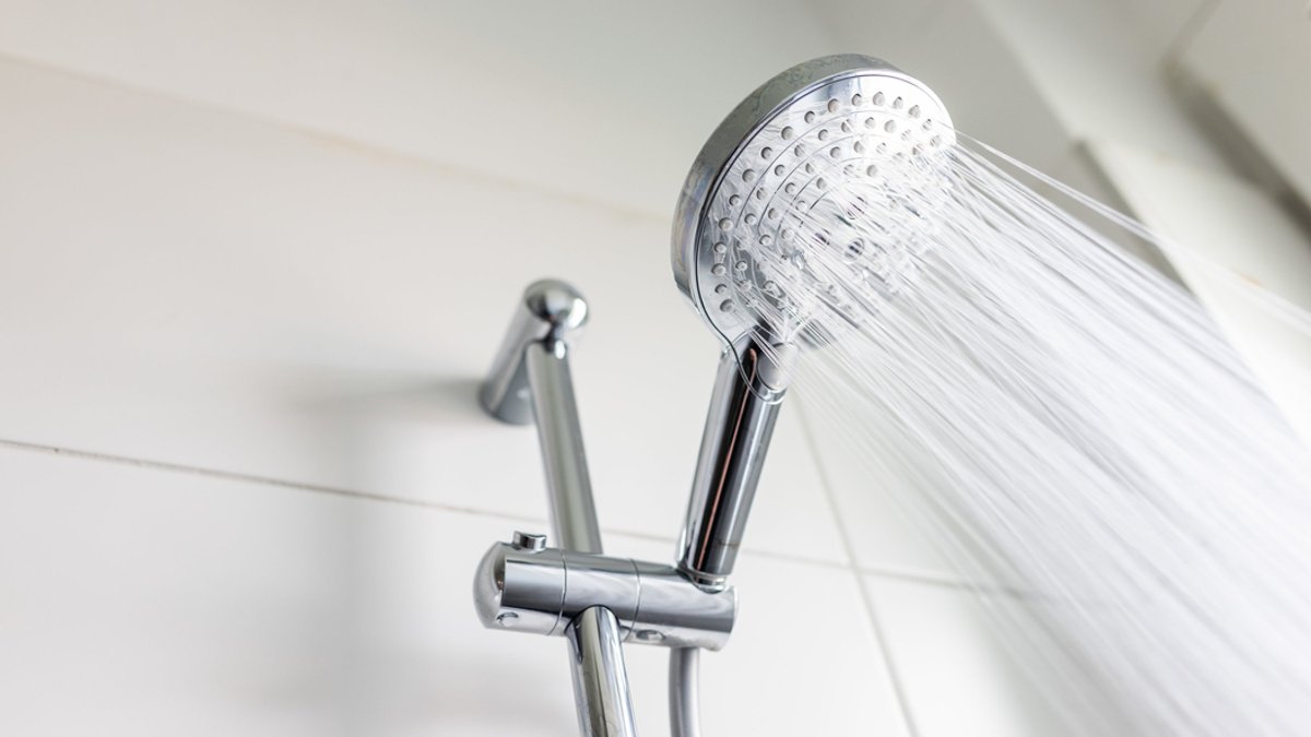 Symbolbild: Aus einem sauberen Duschkopf vor weiß-gefliester Wand fließt Wasser mit hohem Druck.