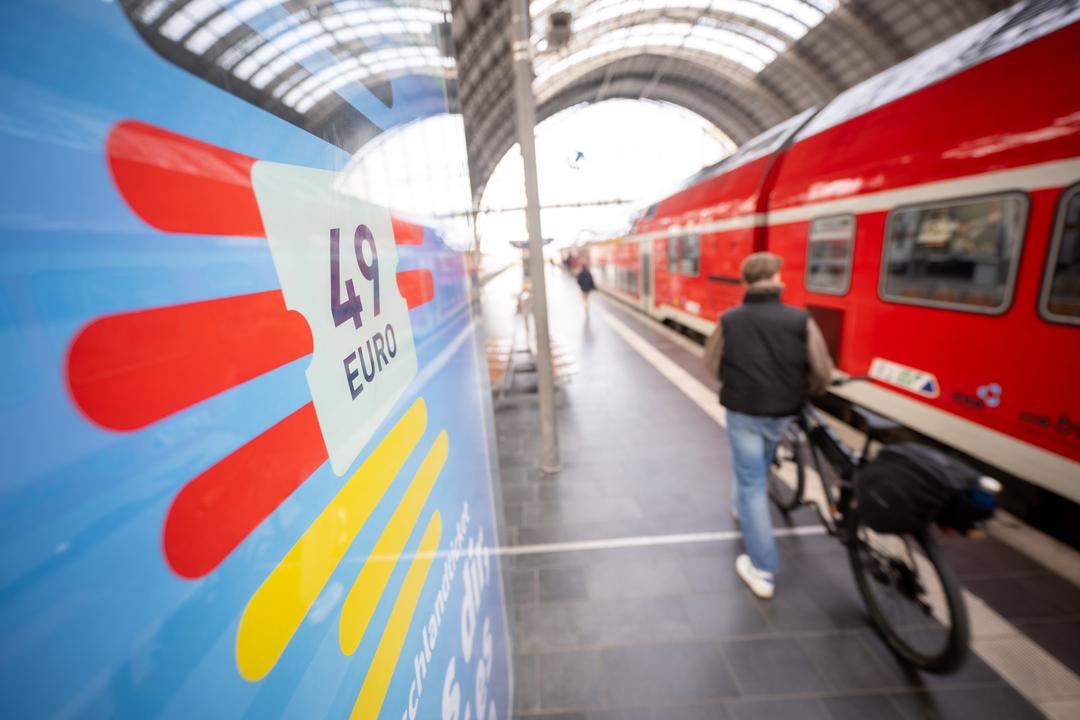 Ein Mann geht am Frankfurter Hauptbahnhof mit seinem Fahrrad zwischen der Werbung für das Deutschlandticket und einer Regionalbahn entlang. 