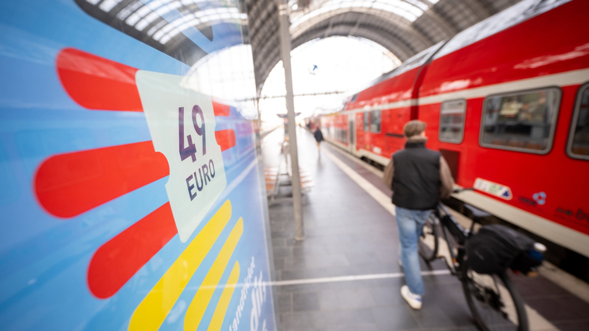 Ein Mann geht am Frankfurter Hauptbahnhof mit seinem Fahrrad zwischen der Werbung für das Deutschlandticket und einer Regionalbahn entlang. 