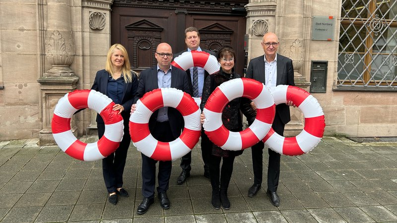Die Kämmerer von Schwabach, Nürnberg, Ansbach, Fürth und Erlangen halten rot-weiß gestreifte Rettungsringe in die Höhe. Sie fordern Hilfe von Bund und Land. | Bild: BR / Tina Wenzel Die Kämmerer von Schwabach, Nürnberg, Ansbach, Fürth und Erlangen halten rot-weiß gestreifte Rettungsringe in die Höhe. Sie fordern Hilfe von Bund und Land.
