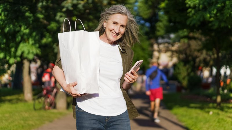 Eine Frau steht mit einer Einkaufstüte unterm Arm und einem Smartphone in der Hand im Park und freut sich. | Bild: picture alliance / Zoonar/svyatoslav lipinskiy Eine Frau steht mit einer Einkaufstüte unterm Arm und einem Smartphone in der Hand im Park und freut sich.