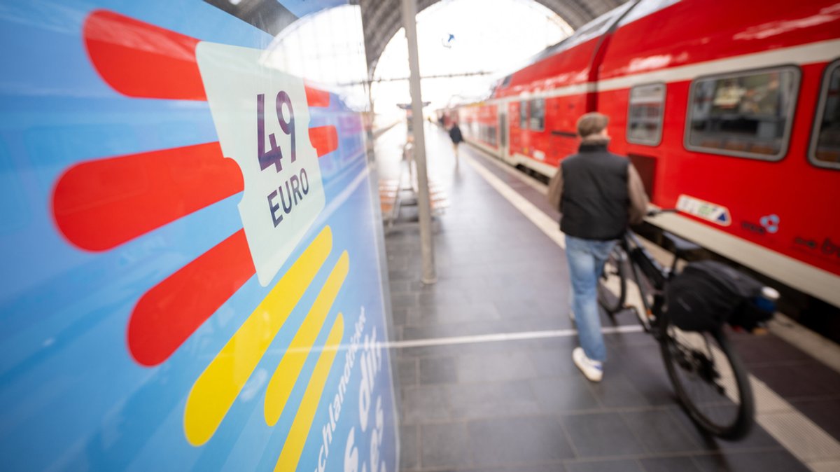 Ein Mann geht am Hauptbahnhof mit seinem Fahrrad zwischen der Werbung für das Deutschlandticket und einer Regionalbahn entlang. 